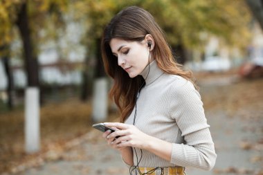 Cep telefonu tutan güzel, şık bir kadın resmi. Genç Avrupalı kız caddede duruyor ve cep telefonu kullanıyor. Kadın telefonla müzik dinliyor ve eğleniyor. Taşınabilir internet konsepti. Sosyal medya.