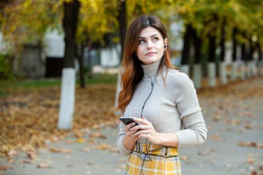 Cep telefonu tutan güzel, şık bir kadın resmi. Genç Avrupalı kız caddede duruyor ve cep telefonu kullanıyor. Kadın telefonla müzik dinliyor ve eğleniyor. Taşınabilir internet konsepti. Sosyal medya.