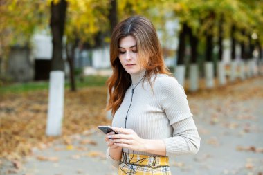 Cep telefonu tutan güzel, şık bir kadın resmi. Genç Avrupalı kız caddede duruyor ve cep telefonu kullanıyor. Kadın telefonla müzik dinliyor ve eğleniyor. Taşınabilir internet konsepti. Sosyal medya.