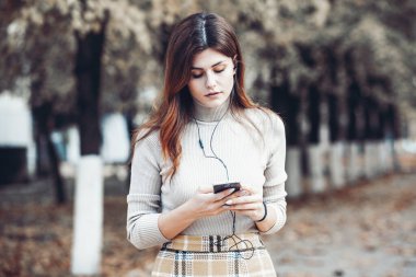 Cep telefonu tutan güzel, şık bir kadın resmi. Genç Avrupalı kız caddede duruyor ve cep telefonu kullanıyor. Kadın telefonla müzik dinliyor ve eğleniyor. Taşınabilir internet konsepti. Sosyal medya.