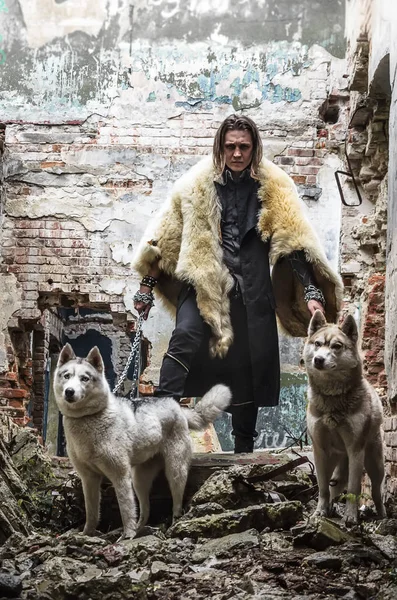 A young man in a leather cloak and a fur cape hides with two dogs — Stock Photo, Image