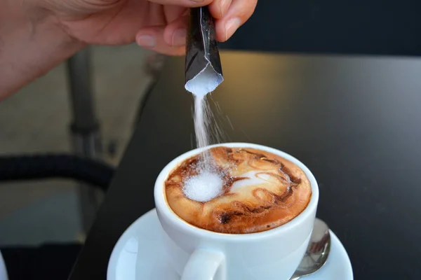 Mano vertiendo azúcar en capuchino caliente . — Foto de Stock