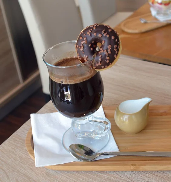 Caffè nero con latte su una tazza di vetro e ciambella al cioccolato su un tavolo di legno . — Foto Stock