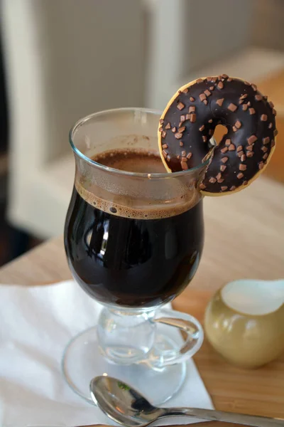 Caffè nero con latte su una tazza di vetro e ciambella al cioccolato su un tavolo di legno . — Foto Stock