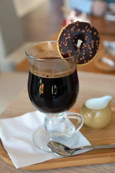 Caffè nero con latte su una tazza di vetro e ciambella al cioccolato su un tavolo di legno . — Foto Stock