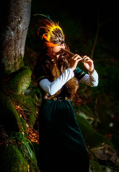 戴着萨满式羽毛面罩 身着古装 在林地周围弹奏木制装饰长笛的女孩 — 图库照片