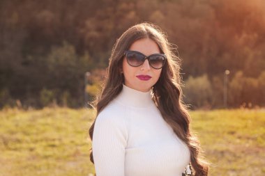 Uzun saçlı ve güneş gözlüklü güzel esmer kadının dışarıda çekilmiş bir portresi. Kameraya bakıyor..