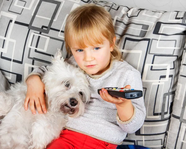 Jovencita rubia viendo la televisión en el sofá con el perrito schnauzer blanco. Niños en aislamiento, cuarentena . — Foto de Stock