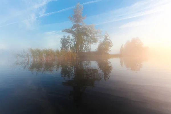 Landscape of morning nature on lake with blue sky, clouds,  fog (mist) and lonely tiny island with trees reflected on the water surface.