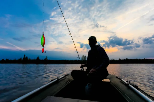 Fisherman silhouette with fishing rod is sitting in the inflatable boat and bright lure on the foreground at sunrise — Stock Photo, Image