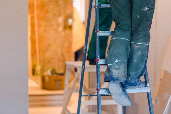 Benen van werknemer met groene werkkleding op de ladder. Werknemer maakt gipsplaten gipsplaat voor gips wanden in het appartement is in aanbouw, verbouwing, renovatie, restauratie en wederopbouw. — Stockfoto