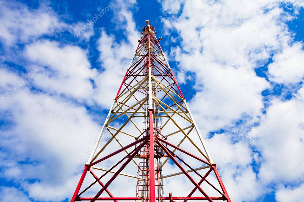 Telecommunication tower with panel antennas and radio antennas and satellite dishes for mobile communications 2G, 3G, 4G, 5G with red fence around tower against blue sky with clouds