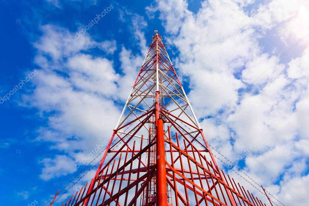 Telecommunication tower with panel antennas and radio antennas and satellite dishes for mobile communications 2G, 3G, 4G, 5G with red fence around tower against blue sky with clouds