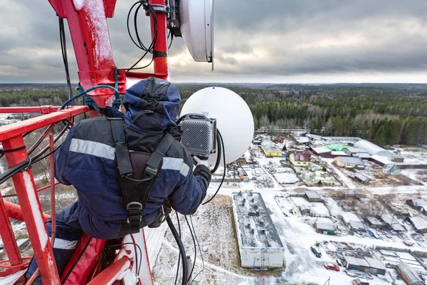 Alpinista industrial profissional em equipamento uniforme e alpinista na torre de telecomunicações está instalando unidade ao ar livre para antena para o próximo ajuste. Processo de trabalho de modernização — Fotografia de Stock