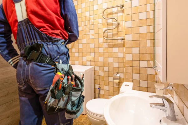 Trabalhador da construção civil e encanador com workwear e saco de ferramentas com ferramentas na cintura permanece banheiro no apartamento para reparar a torneira — Fotografia de Stock