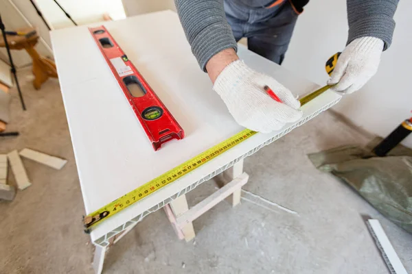 Работник в защитных перчатках измеряет расстояние на строительном материале перед резкой в строящейся квартире, реконструкцией, ремонтом, капитальным ремонтом, расширением, реставрацией и — стоковое фото