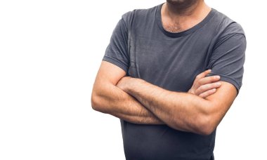 Gri solgun eski moda tişörtlü olgun bir adam beyaz arka planda çapraz kollarıyla duruyor.