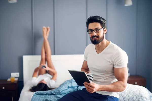 强壮的肌肉男子喝咖啡 并使用平板电脑坐在床上 他的女朋友躺在床上 腿抬高 — 图库照片
