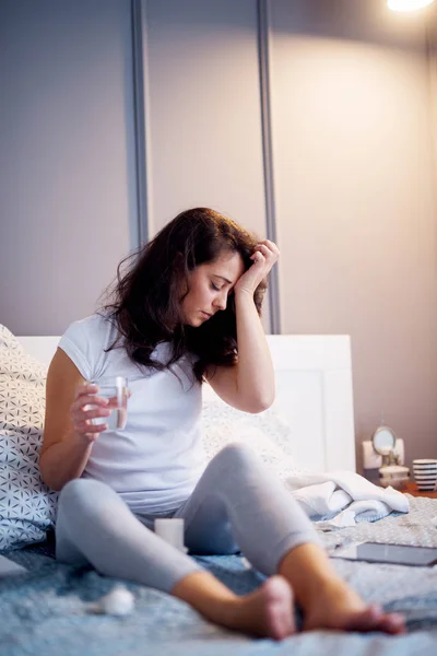 生病的妇女坐在家里的床上 患头痛和饮用水 — 图库照片