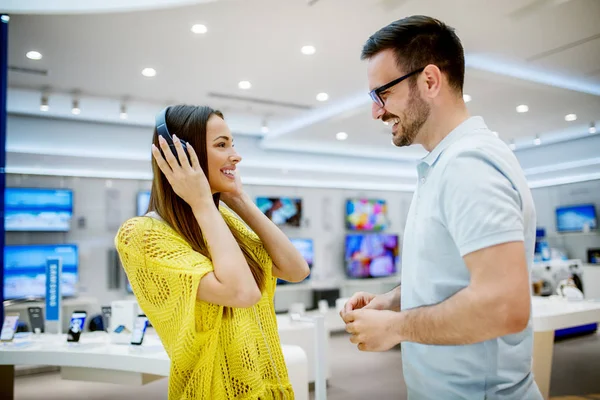 年轻的快乐的女人和男人在技术商店选择新的耳机听音乐 — 图库照片