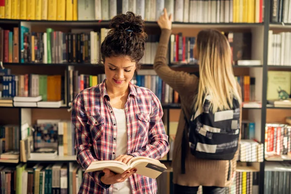 図書館 学生の概念 本棚から本を選択する別の女子学生の前で図書館の本の側に立って幸せな若い女 — ストック写真