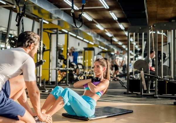 attractive slim woman doing abs exercise on mat with crossed hands with her muscular personal trainer in gym