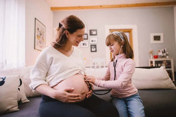 可爱的学步女孩通过听诊器听她怀孕的母亲的肚子 — 图库照片