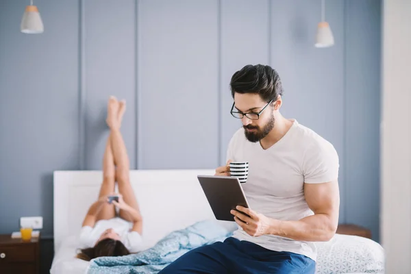 强壮的肌肉男子喝咖啡 并使用平板电脑坐在床上 他的女朋友躺在床上 腿抬高 — 图库照片