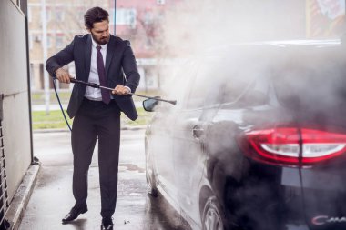 handsome stylish young bearded man in suit washing car at the manual car washing self-service station clipart