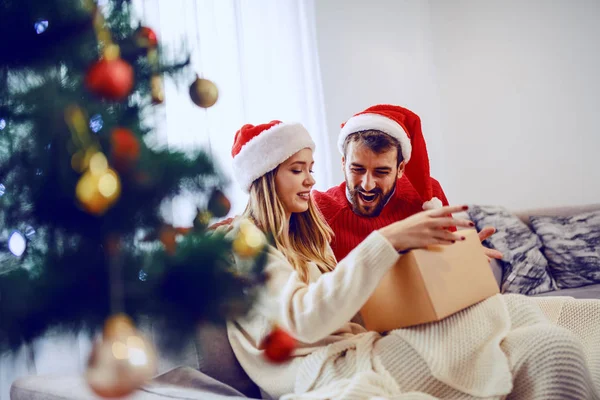 可爱的 面带微笑的夫妇 头戴圣诞礼帽 坐在客厅的沙发上 打开礼物 在前景中的是圣诞树 — 图库照片