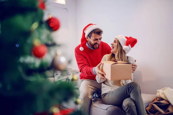 令人惊讶的可爱的白人金发女人坐在客厅的沙发上 接受男朋友的礼物 两个头上都戴着桑塔帽 前景光明的是圣诞树 客厅内部 — 图库照片