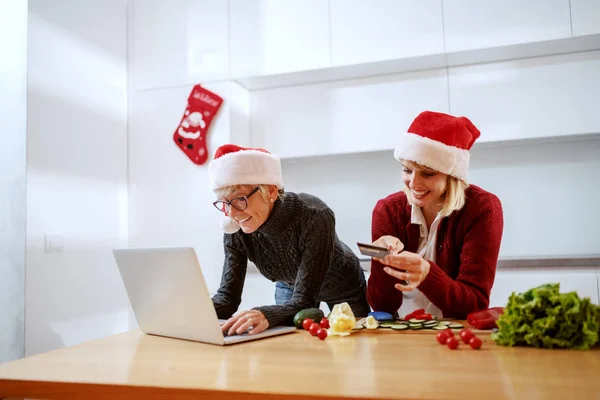 幸せな女性の台所のカウンターにもたれて クレジットカードを見て ノートパソコンに入力する母親 どちらも頭にサンタの帽子を持っている クリスマスショッピングの時間 — ストック写真