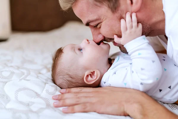 有趣可爱的高加索男孩六个月大的时候躺在床上咬着他爸爸的鼻子 爸爸抱着孩子笑着 — 图库照片