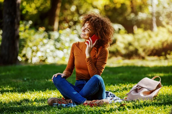 迷人的年轻 面带微笑的混血女子穿着休闲装 卷曲的头发坐在草坪上 在智能手机上聊天 秋天的艳阳天 — 图库照片