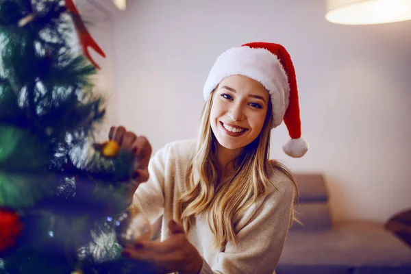 站在客厅里 面带微笑的金发美女头戴圣诞礼帽 看着相机 装饰着圣诞树 — 图库照片