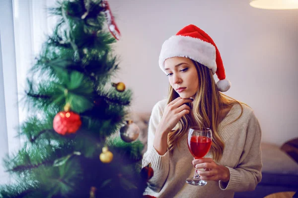 매력적 스웨터를 모자를 머리에 와인을 마시고 거실에 크리스마스 트리를 바라보고 — 스톡 사진