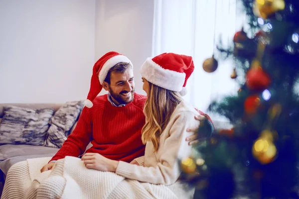 可爱的 快乐的高加索夫妇 头上戴着圣诞礼帽 坐在客厅的沙发上 笑着聊天 在前景中的是圣诞树 — 图库照片