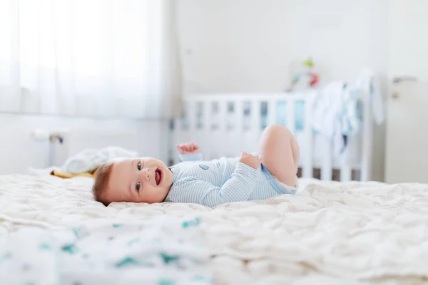 Side view of adorable chubby six months old baby boy lying on bed, smiling and looking at camera. Bedroom interior. Happy childhood concept.