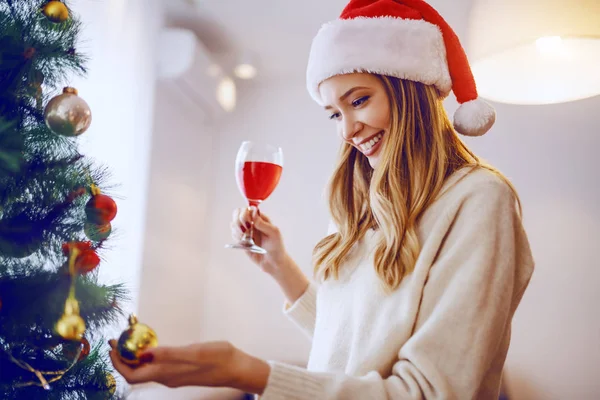 豪華な笑顔Caucasianブロンド女性とともにサンタ帽子でタッチオーナメント上のクリスマスツリーと飲酒ワインながらリビングルームに立って — ストック写真