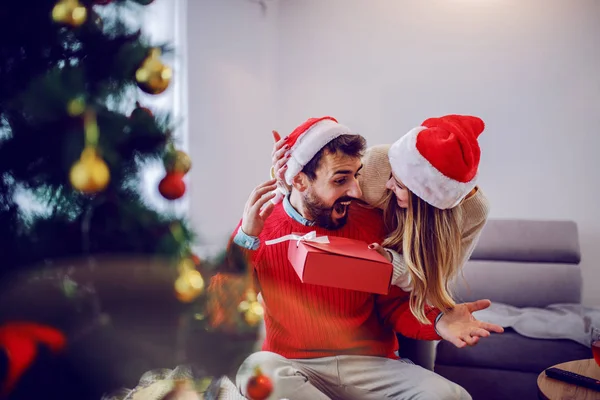 迷人的高加索女人送给她男朋友的圣诞礼物 两人都穿着毛衣 头上戴着桑塔帽 客厅内部 圣诞节假期的概念 — 图库照片