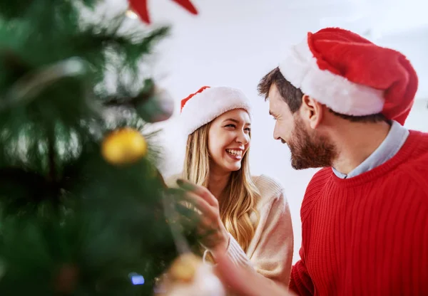 可爱的白种人夫妇装饰着圣诞树 互相望着对方 两个头上都戴着桑塔帽 客厅内部 — 图库照片