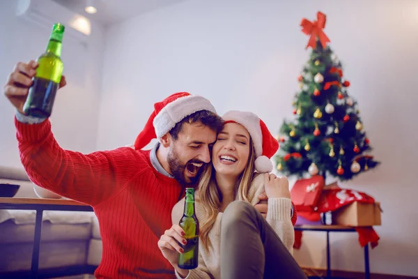 床に座ってビールボトルを持ち 新年を祝う頭の上にサンタの帽子を持つ陽気な白人カップル 背景にはクリスマスツリー リビングルーム内装 — ストック写真