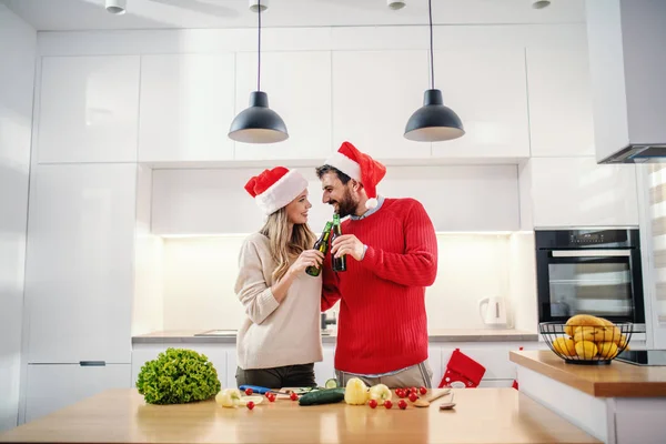 平安夜 一对快乐的白种人夫妇 头戴圣诞礼帽 站在厨房里品酒 厨房柜台上有蔬菜 在前景中的是圣诞树 — 图库照片