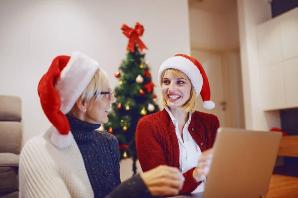 美しい白人ブロンドの女性は リビングルームとラップトップを使用して床の上に母親と座っています どちらも頭にサンタの帽子を持っている 背景にはクリスマスツリーが飾られています — ストック写真