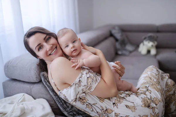 Happy Caucasian mom holding her adorable son while sitting on sofa. Baby looking at camera.