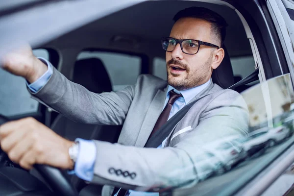 Nervous handsome caucasian businessman in suit and with eyeglasses commenting traffic while sitting in his car. Hand is on steering wheel.