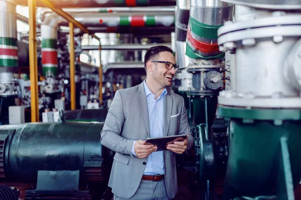 Smiling caucasian supervisor in suit holding tablet and checking on machinery in energy plant.