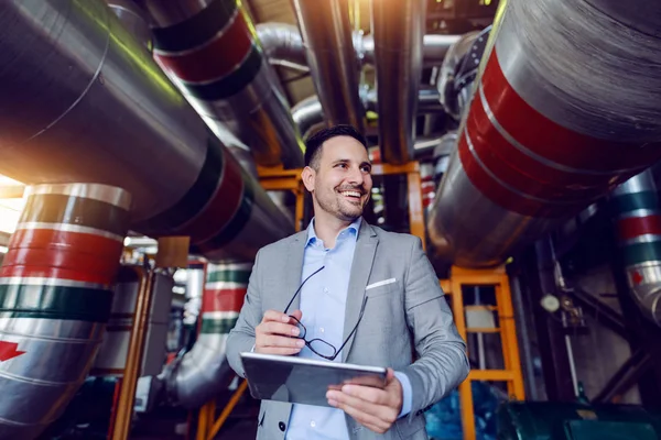 Smiling caucasian supervisor in gray suit holding tablet and eyeglasses in hands and looking away while standing in energy plant.