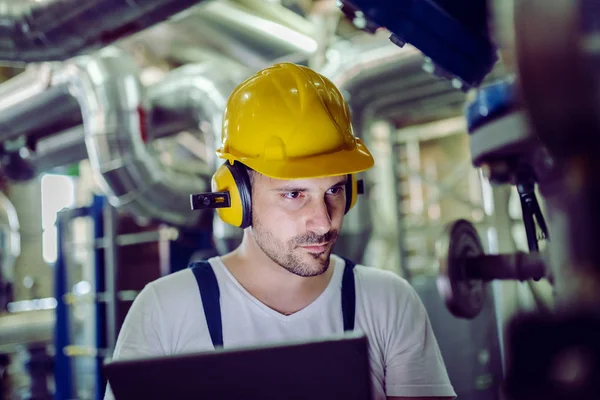 身着工作服的集中工厂工人 头部有防护头盔 耳朵上戴着防磷 使用平板电脑检查机器 — 图库照片