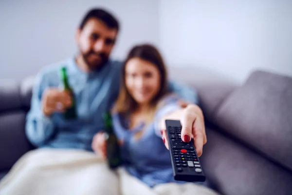 リビングルームにある毛布で覆われた魅力的な白人のカップルは テレビを見てビールを飲んでいます リモコンを持ってチャンネルを変える女 リモートコントロールに焦点を当てる — ストック写真
